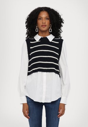 Vest tricotat de culoare bleumarin cu dungi albe orizontale, purtat peste o cămașă albă cu guler, asortat cu blugi clari. Textură netedă și design scurt.