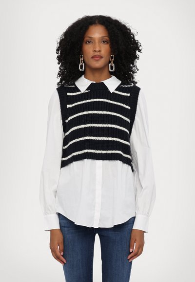 Vest tricotat de culoare bleumarin cu dungi albe orizontale, purtat peste o cămașă albă cu guler, asortat cu blugi clari. Textură netedă și design scurt.
