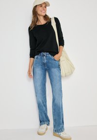 Schwarzes langärmliges Oberteil, blaue gerade Jeans, beiger Sneakers und eine cremefarbene Tasche. Hellbraune Baseballkappe. Einfarbiger Hintergrund.