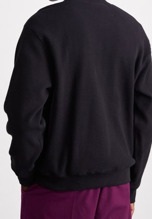 Person wearing a black textured long-sleeve sweater and purple pants, shown from the back with hands in pockets.