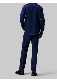 Tmavě modrá džínová košile s zakřiveným spodním lemem, zapínáním na knoflíky na manžetách a tmavě modré džínové kalhoty. Černé boty doplňují outfit. Pohled zezadu.