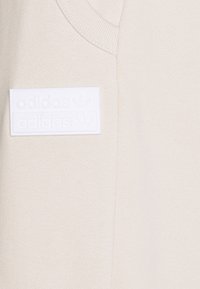 Ljusbeige stickat tyg med en vit rektangulär Adidas-logotypmärke med präglad text och treklöver-symboler på ett plagg med V-ringning.