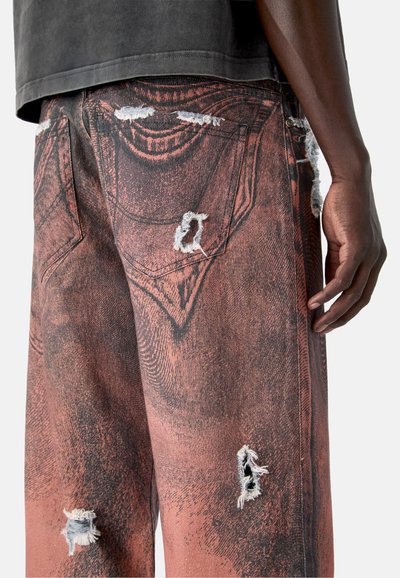 Close-up van een persoon die beschadigde roodbruine korte broek draagt met bedrukt zakdetail en rafelige gaten, staand tegen een effen achtergrond.