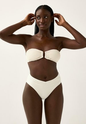 Nainen, jonka hiukset on punottu, säätää olkaimettoman, teksturoidun valkoisen bikinitopin ja korkeavyötäröiset alaosat yksinkertaista vaaleaa taustaa vasten.