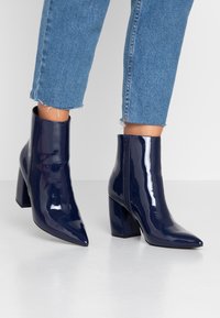 Botas de tobillo de charol azul marino con puntera puntiaguda, tacón alto tipo bloque y cremallera lateral, combinadas con unos jeans de mezclilla recortados y deshilachados.