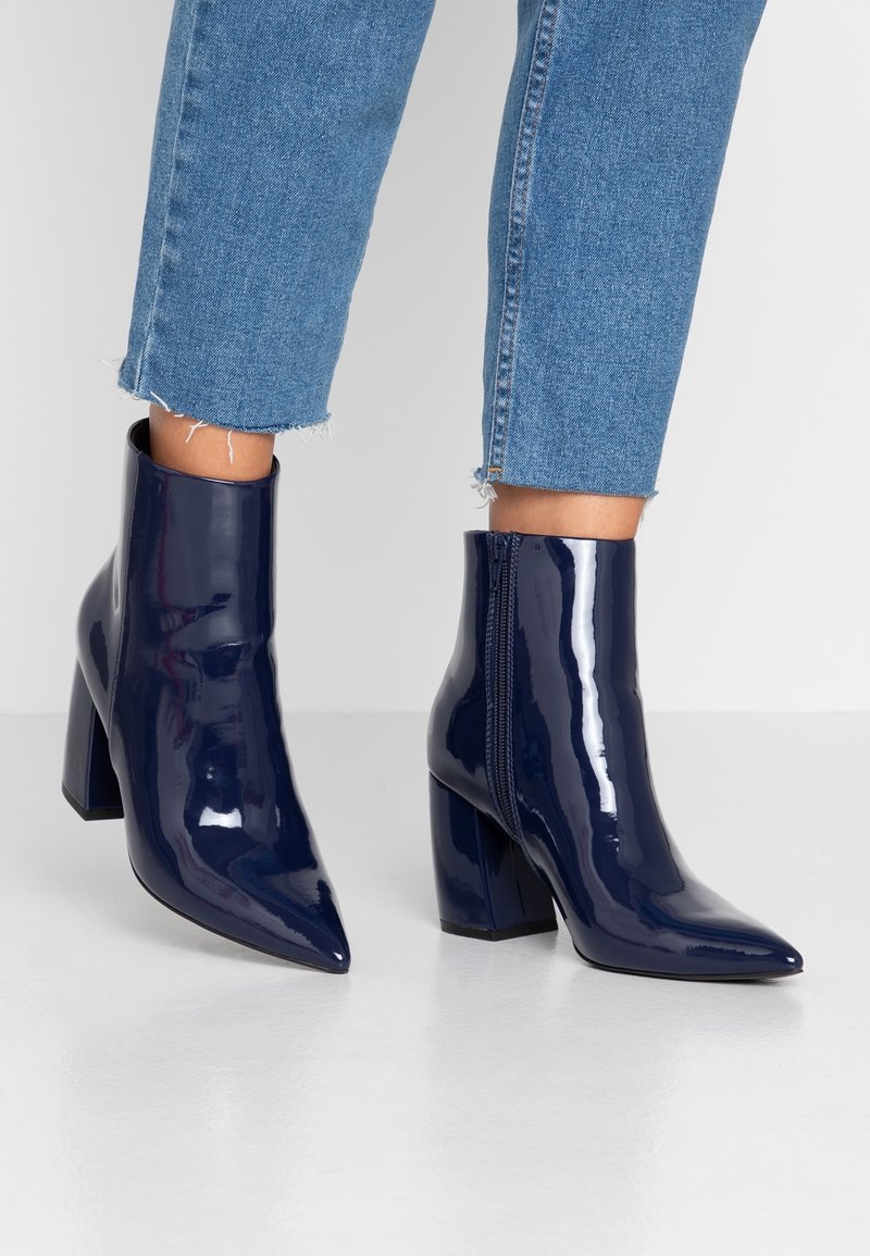 Botas de tobillo de charol azul marino con puntera puntiaguda, tacón alto tipo bloque y cremallera lateral, combinadas con unos jeans de mezclilla recortados y deshilachados.