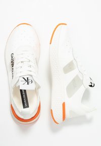 White Calvin Klein sneakers with orange accents, mesh panels, and black branding on the sides and tongue, shown from top and side views.
