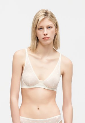 Ξανθιά γυναίκα με κοντά μαλλιά φοράει ένα διάφανο λευκό σουτιέν και ταιριαστά εσώρουχα με χαμηλή μέση, σε απλό ανοιχτό φόντο.