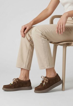 Osoba siedząca na beżowym krześle, ubrana w beżowe spodnie i brązowe mokasyny zamszowe z ozdobami z koralików i beżowymi sznurowadłami.