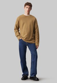 Camisa marrón texturada de manga larga con cuello redondo, combinada con jeans rectos azul y zapatos negros. Diseño sencillo y casual.