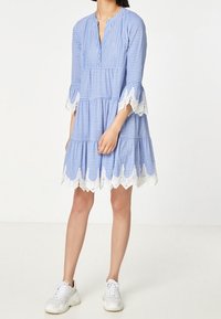 Modro-bílé pruhované šaty s volnější siluetou, zdobené krajkovými detaily na rukávech a spodním lemu, v kombinaci s bílými teniskami.