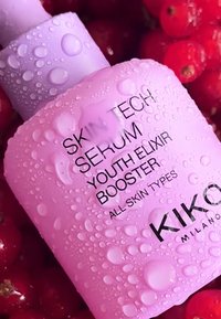 Rosa serumflaske med en teksturert overflate dekket av vanndråper. Har svart tekst med produktdetaljer, plassert mot en bakgrunn av røde bær.