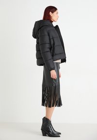 Doudoune noire avec capuche et texture matelassée, associée à une jupe en cuir noir avec des franges et des bottines pointues.