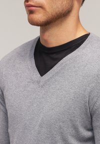 Pull gris à col en V, en tissu tricoté doux, finition texturée, coupe ajustée, superposé à un t-shirt noir à col rond.