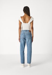 Mujer con cabello trenzado que lleva una blusa blanca off-shoulder, jeans de cintura alta de color azul claro y zapatillas blancas, de pie frente a un fondo neutro.