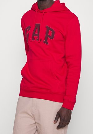 Mand iført en skrigrød GAP-hættetrøje med stor logo-tekst, kombineret med lysebeige bukser, stående mod en ensartet lys baggrund.