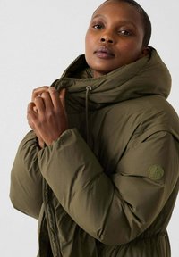 Chaqueta acolchada color verde oliva con cuello alto y capucha ajustable. Presenta un parche con el logo en la manga y una cintura ceñida para un mejor ajuste.