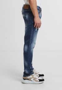 Slim fit modré denimové džíny s bledým ombré, drobnými oděrkami a hnědou koženou nášivkou na pase, v kombinaci s vícebarevnými teniskami.