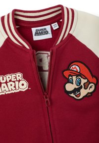 Chaqueta roja y crema con el logo de Super Mario y la cara de Mario bordada, parcialmente desabrochada, mostrando la etiqueta de tamaño para 4-5 años.