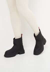 Botines negros hasta el tobillo con un acabado mate suave, suela de goma gruesa, punta redonda y una tira en la parte trasera que presenta detalles en rojo y blanco.