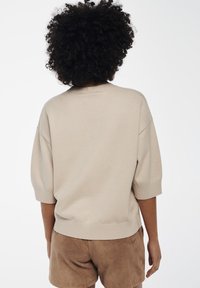 Beige gebreide trui met korte mouwen en ribbelboord. Draagt met bruine suède shorts, gezien van achteren. Soepele textuur.