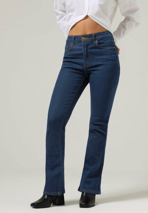 Kvinde iført mørkeblå bootcut-jeans med høj talje, hvid kort top og sorte ankelstøvler med hæl, stående mod en ensfarvet baggrund.