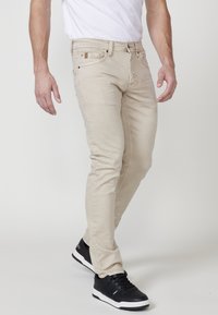 Jeans ajustados beige de mezcla de algodón, con un diseño de cinco bolsillos, cinturillas y cremallera, combinados con zapatillas negras.