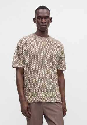 Jeune homme aux cheveux courts portant un pull à manches courtes beige et marron à motif zigzag et un pantalon marron, debout devant un fond blanc.