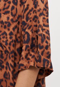 Brun og svart leopardmønstret skjorte med korte ermer, brystlomme og knappdetalj på ermene for å kunne rulle dem opp. Glatt tekstur.
