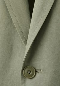 Gros plan sur le revers d'un blazer vert olive avec des coutures apparentes et un gros bouton rond vert-brun fixé dans la boutonnière.