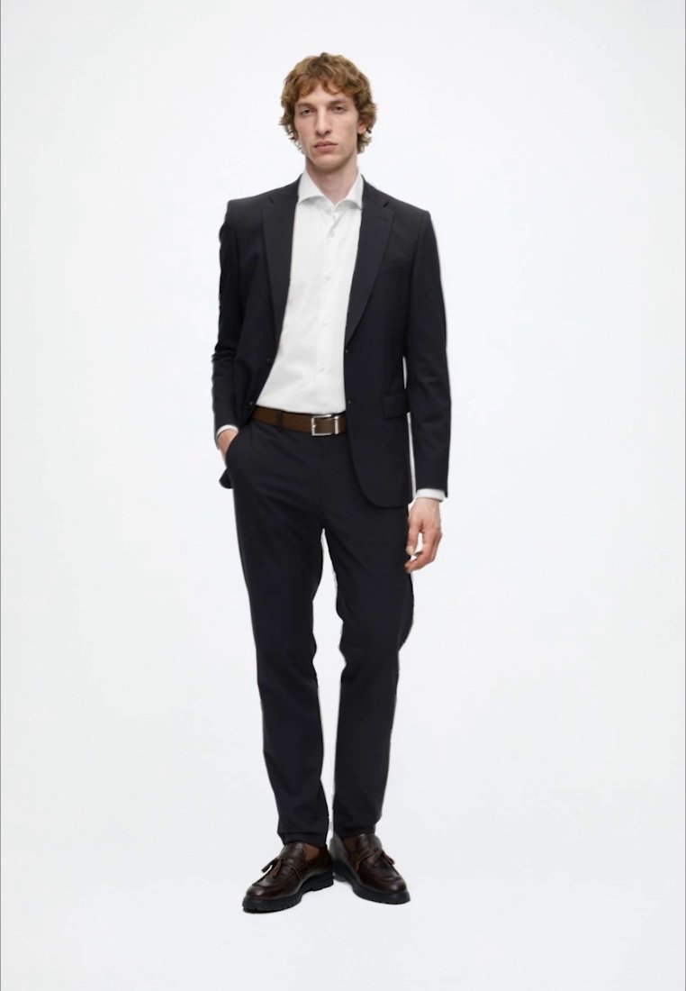 Jeune homme debout portant un costume noir, une chemise blanche, une ceinture marron et des chaussures marron foncé sur un fond blanc uni.