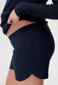 Granatowe szorty z prążkowanej tkaniny z falbaniastym dołem, z elastycznym paskiem w talii i gładką fakturą, pokazane z boku z delikatnie uniesioną bluzką.