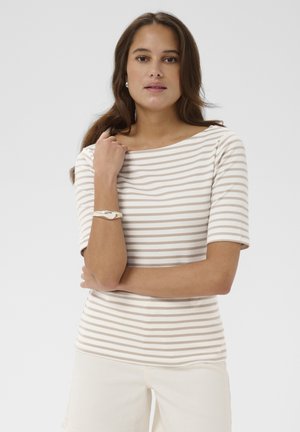 Femme aux cheveux bruns portant un haut à manches courtes beige et blanc à rayures, un pantalon écru, un bracelet argenté et des boucles d'oreilles, posant devant un fond uni.