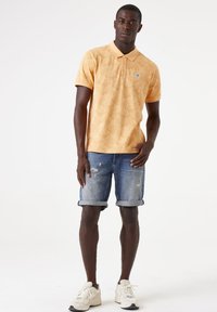 Camisa de polo naranja con estampado y cuello, combinada con pantalones cortos de mezclilla desgastados y doblados en el dobladillo, y zapatillas blancas.