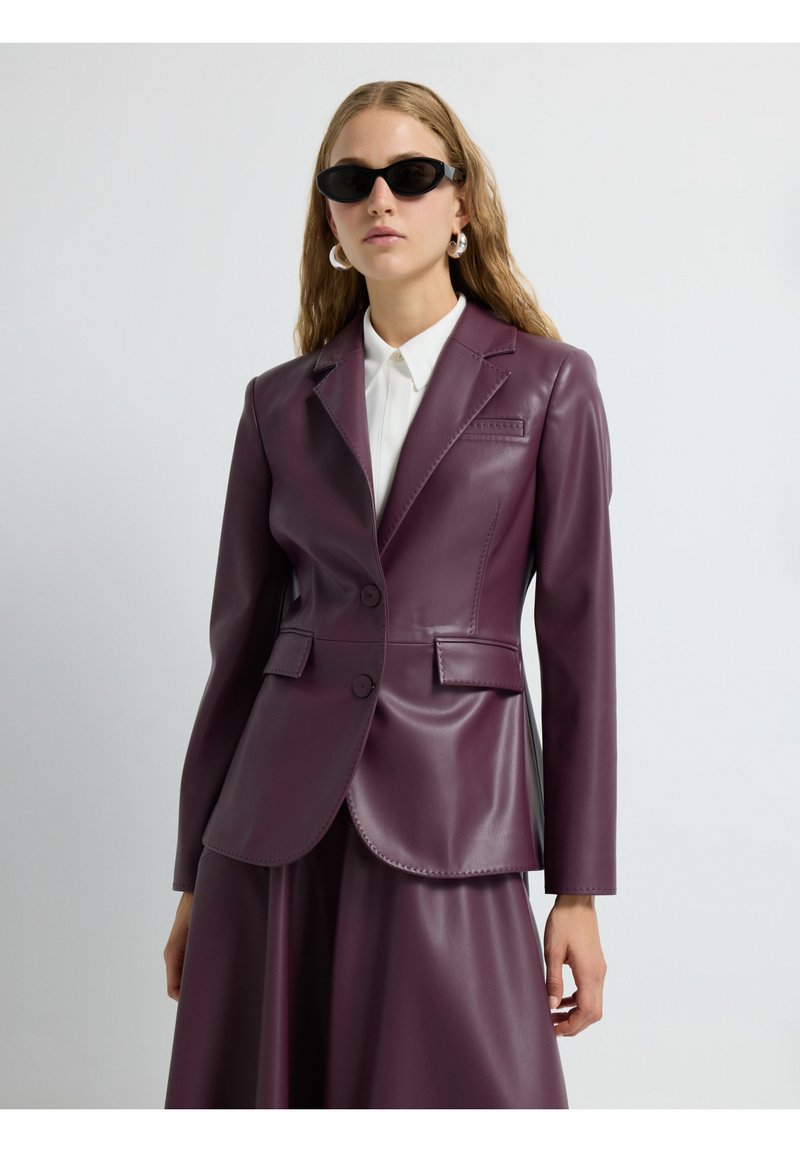 Blazer in finta pelle borgogna con due bottoni, revers a punta, tasche laterali e una texture liscia e lucida. Camicia bianca con colletto sotto.