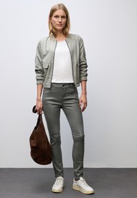 Jonge vrouw in een lichtgrijze bomberjack, witte t-shirt, olijfgroene broek, witte sneakers en een bruine suède handtas tegen een effen achtergrond.