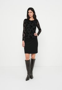 Μαύρη μπλούζα με μακριά μανίκια και λουλουδάτα σχέδια, συνδυασμένη με μαύρη φούστα μέχρι το γόνατο και καφέ ψηλές μυτερές μπότες μέχρι το γόνατο. Ομοιόμορφη υφή, κομψή εφαρμογή.