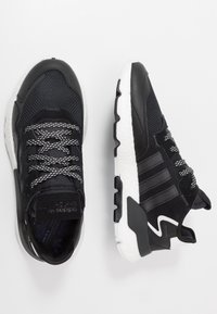 Par av svarta Adidas-sneakers med vita sulor och mönstrade skosnören, som visar topp- och sidovy på en vit bakgrund.