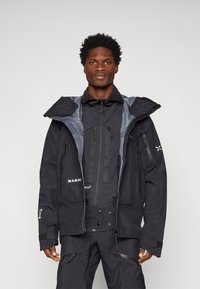 Casaco preto à prova de água com detalhes cinzentos, capuz grande, múltiplos bolsos com zíper e punhos largos, sobre uma roupa escura.