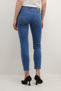 Jeans en denim bleu avec une coupe skinny, taille intermédiaire et poches arrière visibles ; associés à des chaussures à talons hauts noires sur un fond neutre.