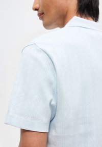 Άνδρας που φοράει ανοιχτό μπλε πουκάμισο με κοντά μανίκια και διακριτικά κάθετα ζιγκ-ζαγκ και τεθλασμένα σχέδια, απεικονίζεται από τον ώμο μέχρι τον λαιμό.