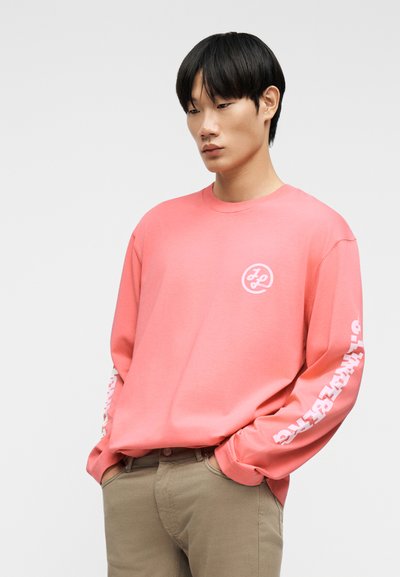 Νέος άνδρας με μαύρα μαλλιά, φορώντας κοραλί μπλούζα με μακριά μανίκια, λευκό λογότυπο στο στήθος και κείμενο στο δεξί μανίκι, με τα χέρια στις τσέπες χακί παντελονιού.