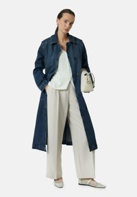 Denim lange jas in donkerblauw, met knoppen en zakken, gecombineerd met een lichtbeige top en wijde beige broek, afgewerkt met lichte schoenen.