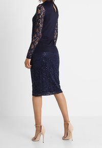 Marineblå klänning med långa spetsärmar, figurnära liv och en knä-length paljettkjol. Bakstycket har en nyckelhål-detalj. Bärs med hudfärgade klackar.