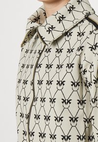 Chaqueta beige con un patrón floral negro, con un gran cuello y detalles de cremallera dorada, que muestra una tela tejida texturizada.