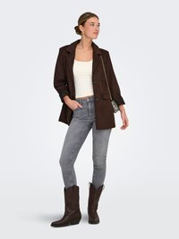 Brown suede blazer with a lapel, paired with a white tank top and gray skinny jeans, complemented by brown cowboy boots and a small bag.