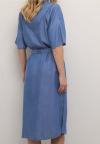 Denim-mekko, jossa on lyhyet hihat, vaaleansininen väri, vyötärölle sidottu vyö ja V-muotoinen takarintakaulus. Polvipituinen, jossa on sivuaukot liikkumisen helpottamiseksi.