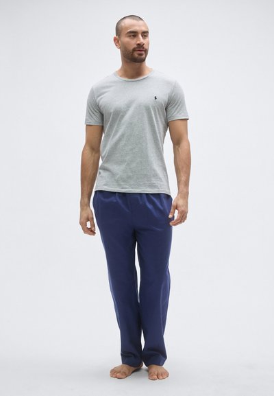 Pelēka kokvilnas T-krekls ar apaļu kakla izgriezumu un nelielu logotipu, kopā ar tumši ziliem, vaļējiem biksēm no viegla materiāla.