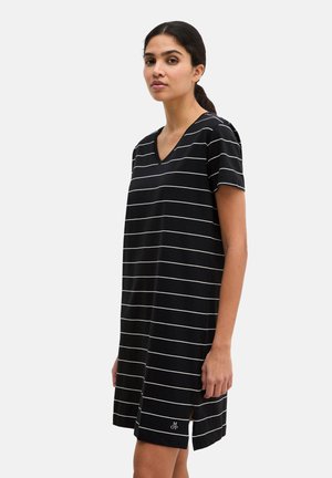 Kobieta ubrana w czarną sukienkę z krótkim rękawem i dekoltem w serek, z białymi poziomymi paskami, stojąca na tle jednolitego, jasnego tła.