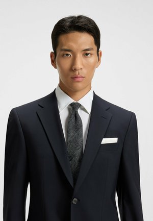Costume bleu marine avec un revers net, chemise habillée blanche et cravate en chevrons texturée. Comprend un mouchoir de poche blanc dans la poche poitrine.